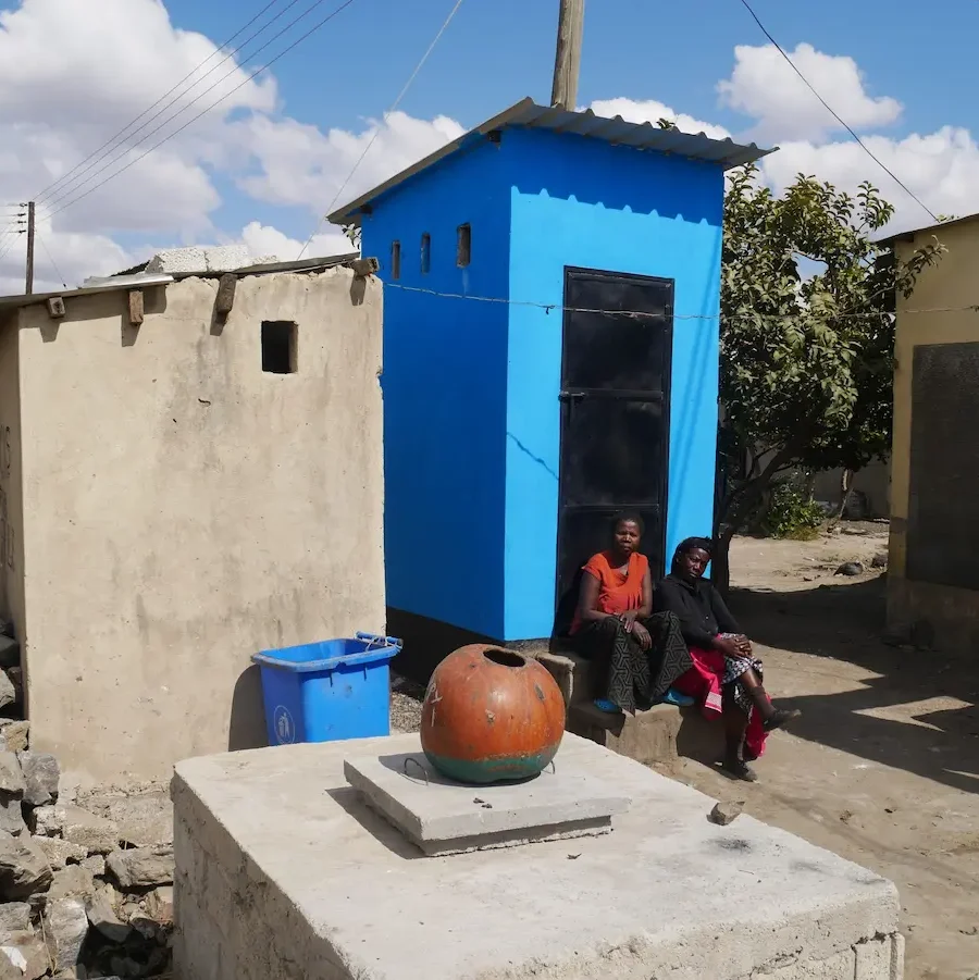 two women sit on the steps of their new blue latrine, next to the old one